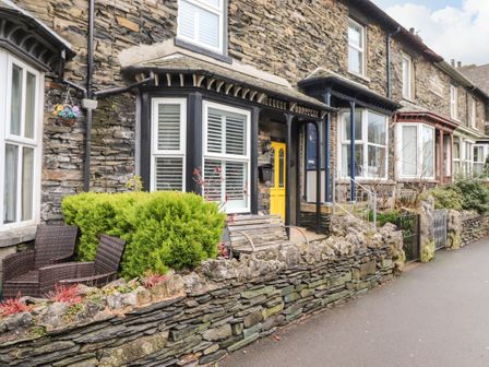 A stone terrace with chairs bushes a wooden bench and a yellow door at Snowflake Cottage in Windermere