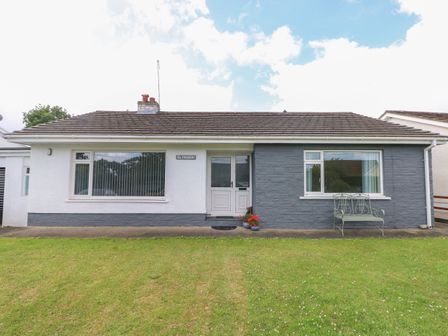 A single story house with white and gray exterior walls a white door two large windows a metal bench and a grass lawn at Glynderi in Cilgerran