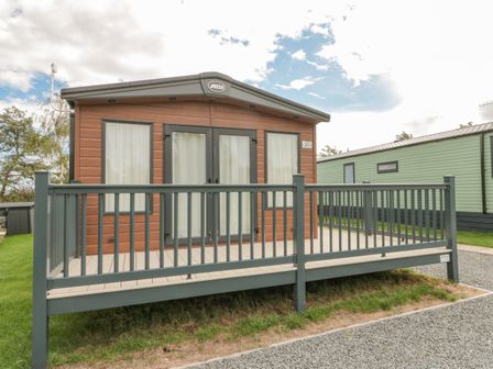 A small cabin with brown siding and black trimmed windows and doors with a gray wooden railing in front at 20 Wolds View in East Heslerton near West Heslerton