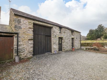 A stone building with wooden doors and windows on a gravel area with trees in the background at Myras View in Newby Bridge