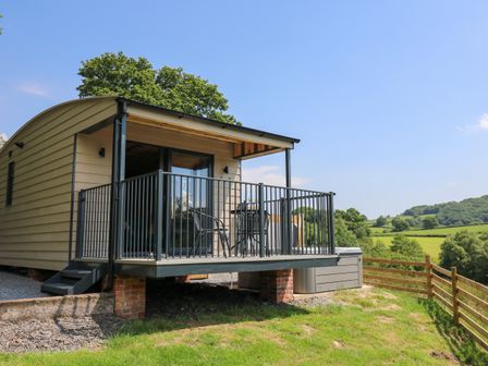 A small raised cabin with a balcony and outdoor chairs overlooking a grassy landscape at Rooster's Rest in Rhaedr Gwy near Rhayader