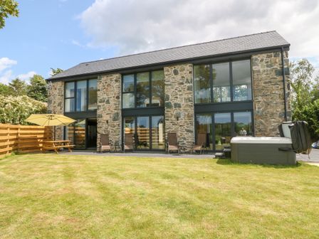 A stone house with large windows and doors a grassy yard with patio chairs a wooden fence and a hot tub at Pen-Y-Bryn in Llanfairpwllgwyngyll