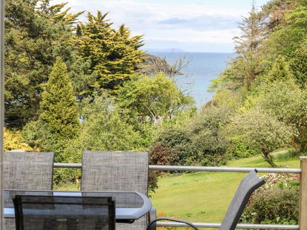 A garden with trees and a sea view behind outdoor chairs and a table at Flushing Meadow in Manaccan near Helford