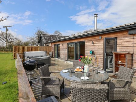 An outdoor patio area with a round glass table and wicker chairs a barbecue grill a hot tub and a wooden building in the background at Strawberry Bank - Nuthatch in Pyworthy near Bridgerule