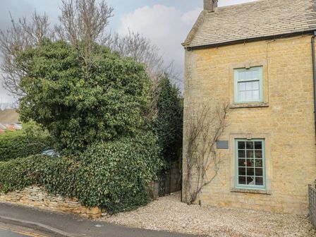 A stone house with two windows next to a gravel path and dense green bushes at Barton Cottage in Bourton-On-The-Water