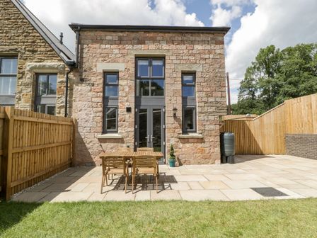 A stone house with a wooden table and chairs on a patio and a grassy lawn at 2 The Old School in Ruspidge