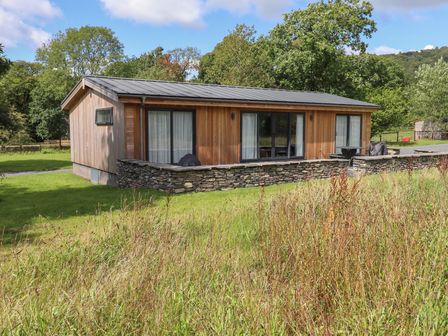 A single-story wooden cabin with stone wall and sliding glass doors surrounded by grass and trees at Corning Mill View in Haverthwaite