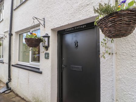 The front entrance of Cobble Cottage with a black door a knocker and two hanging baskets with flowers at Keswick