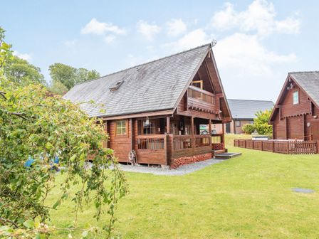 A wooden cabin with a porch and balcony surrounded by grass and trees at Robin Lodge in Llanbedr