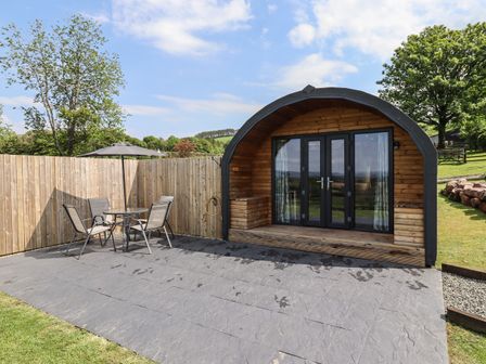 A curved wooden cabin with glass doors on a stone patio with a table umbrella and chairs at Cleugh Lodge in Lockerbie