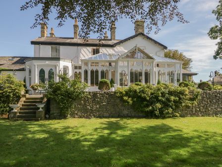 A backyard with grass bushes and stone wall in front of a white house with multiple chimneys and a glass conservatory at Valley View in Dalton-In-Furness