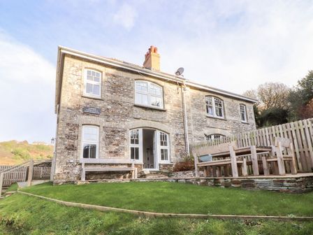 A stone house with white framed windows and a wooden outdoor bench and table set on a raised stone patio at Cliffside Trebarwith Strand near Tintagel