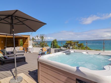 An outdoor deck with a hot tub lounge chairs umbrellas and seating area overlooking the ocean at Tresco Bungalow in St Ives