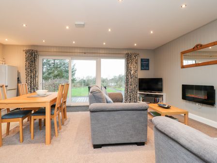 A living room with grey sofas a wooden dining table and chairs a television and sliding glass doors at Beech Lodge in Goosnargh