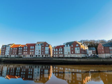 A row of residential buildings reflected in a river with a clear blue sky at Whitby Waves in Whitby