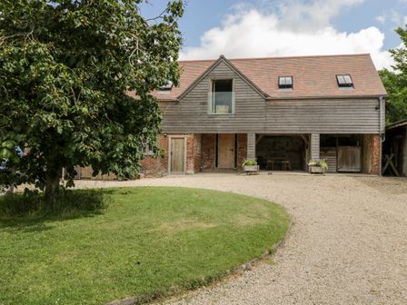 A barn style building with wooden doors and a balcony above a gravel driveway and a tree on the left at The Wool Barn in Upton Lovell near Warminster