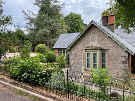 A stone house with a bay window surrounded by a garden and iron fence at Court Lodge in Bathealton near Wiveliscombe