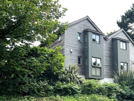A house with gray wooden siding surrounded by green trees and plants at 7 Toms Yard in St Ives