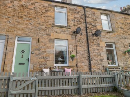An outdoor view of a stone cottage with a green door two windows a small grey fence a small table and two chairs with cushions hanging plants and satellite dishes at Cosy Cottage in Amble
