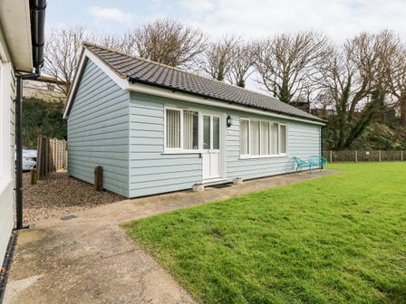 A single-story house with blue exterior siding white door and windows two blue chairs on the patio and a grassy lawn at 1 The Dell in Mundesley