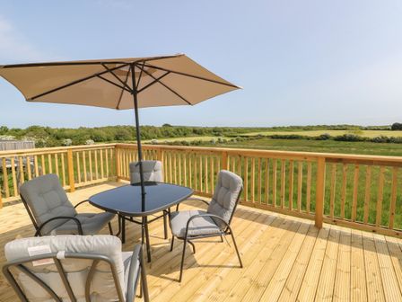 A wooden deck with a table, four cushioned chairs, and a large umbrella overlooking green fields at Mountain View Lodge 2 in Caergeiliog near Valley