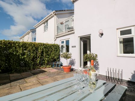 A patio area with a blue picnic table holding a bottle of wine and four wine glasses outside a white house with potted plants at 3 West Park Mews in Hope Cove