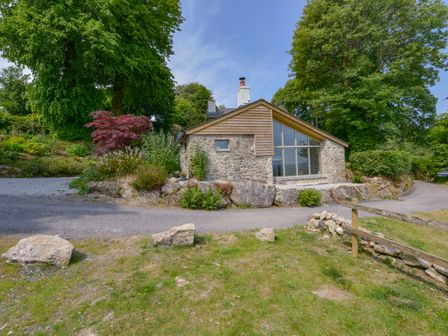 A stone and wood house surrounded by trees and plants beside a gravel road at Beckaford View in Manaton near Ilsington