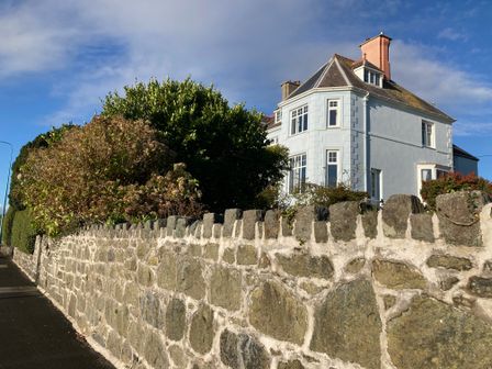 A stone wall with bushes behind it and a blue house with multiple windows in the background at Llys Owen in Criccieth