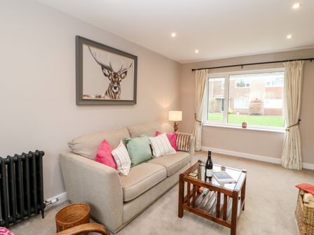 A living room with a beige sofa with colorful cushions a wooden coffee table with a bottle and book and a window with cream curtains at Willows in Corbridge