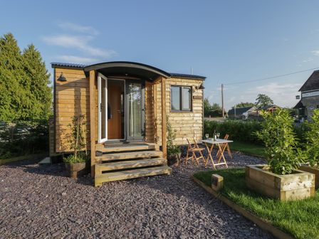 A small wooden cabin with open double doors and steps leading to a gravel area with a table and chairs at Willow in Kemble