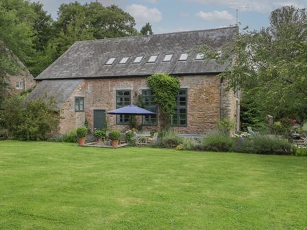 A stone house with multiple windows and a patio with a table umbrella and chairs surrounded by lawn and trees at Owl Barn Marston Stannett near Leominster