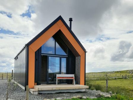 A small modern cabin with orange and black accents and a bench on the porch in a grassy rural area at North Light Studio in Locheport near Lochmaddy North Uist