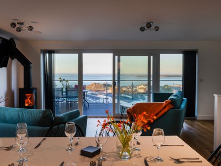 A dining table set with glasses utensils and flowers in a living room with sofas and a fireplace overlooking a balcony and sea view at Lanyon in St Ives