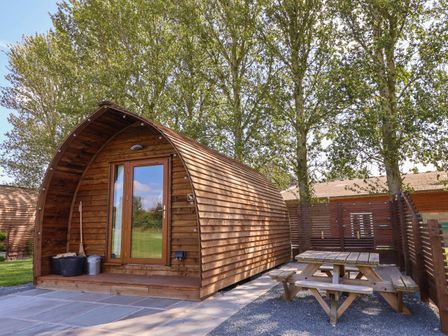 A wooden pod cabin with a glass door next to a wooden picnic table and bench set surrounded by trees at Covey in Little Kelk near Bridlington