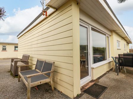 An exterior corner of a single-story building with a sliding glass door two wooden chairs and a table with chairs on a patio at Lapwing in Perranporth