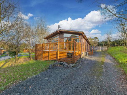 A wooden cabin with a fenced deck and gravel driveway surrounded by trees at Fern Lodge in Allithwaite near Cartmel