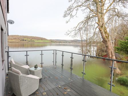 A balcony with two chairs a small table and glass railing overlooking a lake and hills at Vyrnwy Lakeside Bala Lake