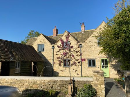 A stone cottage with a green door surrounded by a stone wall and trees at High Cogges Farm Holiday Cottages in Witney