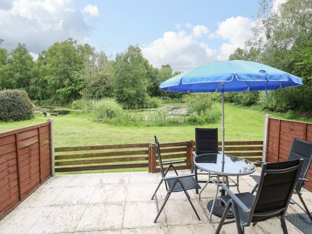 A patio with a glass table four chairs and a blue umbrella overlooking a grassy area and pond at 10 Ruston Reaches East Ruston near Stalham