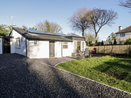 A single-story white house with a gravel driveway and outdoor seating area on a wooden deck at The Cottage at Cromlech Manor Farm in Llanfair-Mathafarn-Eithaf near Benllech