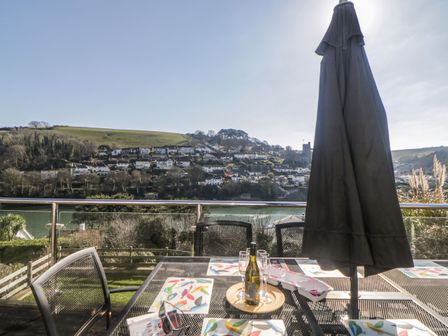 An outdoor patio table with placemats glasses and a bottle of wine with a large closed umbrella and a view of houses and hills at Estuary View in Newton Ferrers