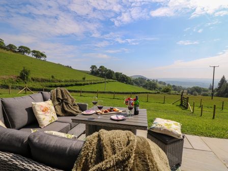 An outdoor seating area with gray cushioned chairs pillows a table with wine glasses and snacks overlooking green fields at Dol Blodau in Newbridge-On-Wye