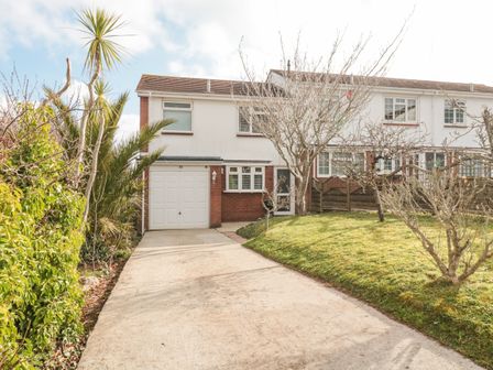 A driveway leading to a garage of a semi detached house with trees and bushes in the front garden at Bay View in Paignton