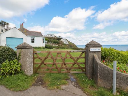 Wooden gate with a concrete fence and pillars opening to a green lawn with houses and a sea view at Beachcliff in Gorran Haven