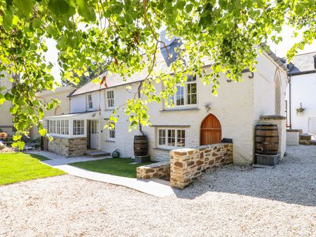 A white stone house with a wooden door and windows surrounded by gravel and grass with barrels near the walls at Trewince Manor Cottage in Portscatho