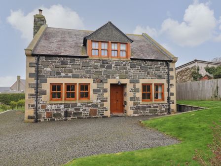 A stone house with wooden framed windows and a wooden door with a gravel driveway and green lawn at Whin Hill Cottage in Craster
