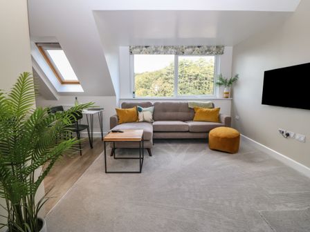 A living room with a gray sofa with yellow cushions a coffee table a round yellow ottoman a dining table with chairs and a large window at Apartment 16 in Y Felinheli