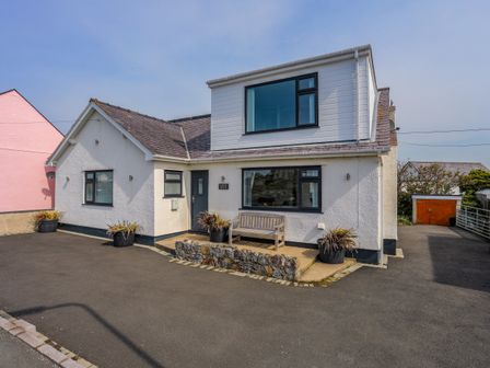 A white house with a grey door bench and planters outside on a paved area at Porth Ewyn in Trearddur Bay