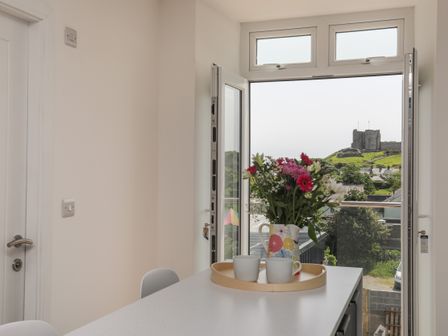 A kitchen table with flowers in a vase and two mugs near an open door with a view of a castle on a hill at Swn Y Mor Sound of the Sea in Criccieth