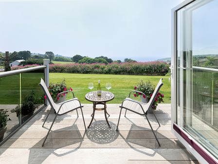 A balcony with two chairs and a small table with a bottle and two glasses overlooking a garden and distant hills at Greenside Studio in Praa Sands